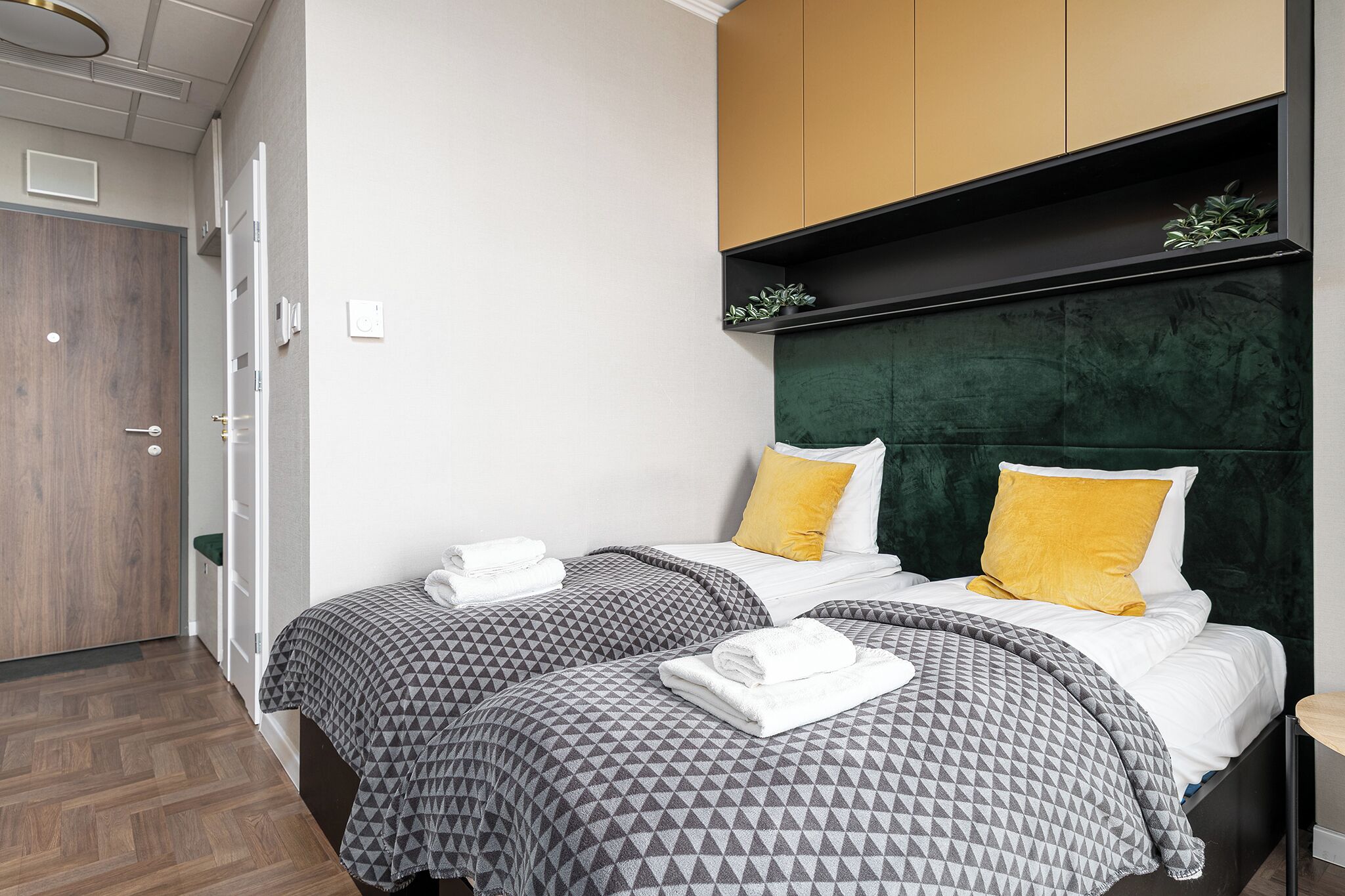 A hotel room with two beds, each with patterned bedding and a green headboard. There is a wooden door and a small plant on a shelf above the beds.