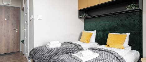 A hotel room with two beds, each with patterned bedding and a green headboard. There is a wooden door and a small plant on a shelf above the beds.