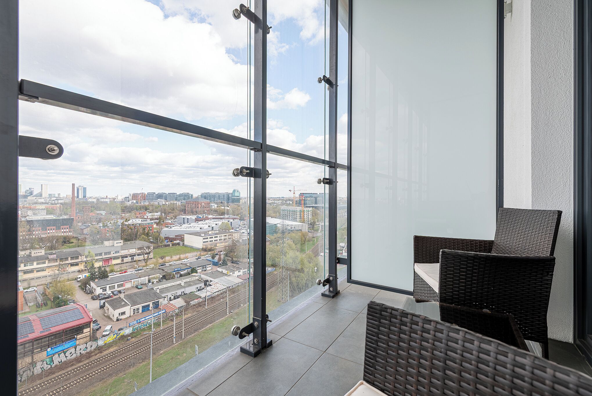 A balcony with a city view, a wicker chair, and a glass railing.