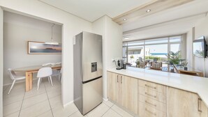 A modern kitchen with a stainless steel refrigerator, wooden cabinets, a dining area with a table and chairs, and a large window offering a view of the outdoors. - Dolphin Beach CG12, Blouberg by HMH (Cape Town)