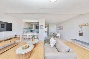 A modern living room with a sofa, coffee table, and TV. There is a kitchen in the background with a dining area.