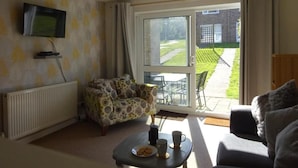 Un salon avec un canapé, une table basse sur laquelle se trouvent des tasses et une assiette, un téléviseur fixé au mur et une porte-fenêtre coulissante donnant sur un jardin.