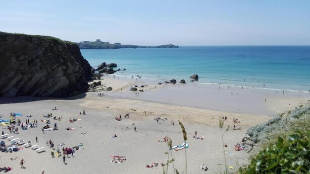 Une plage où des gens se prélassent au soleil et se baignent, une falaise rocheuse sur la gauche et une mer bleue et limpide.