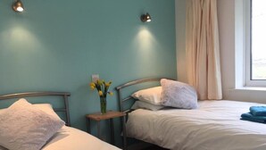 Une chambre avec deux lits, chacun étant équipé de draps blancs et d’une serviette bleue. Une petite table avec un vase de fleurs jaunes se trouve entre les deux lits.