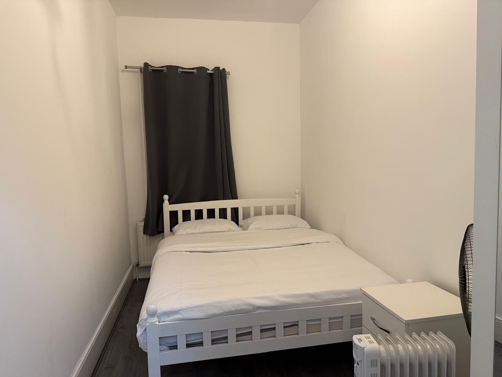 A small, white-framed bed with a white mattress and pillows, a black curtain, a white bedside table, and a white radiator.