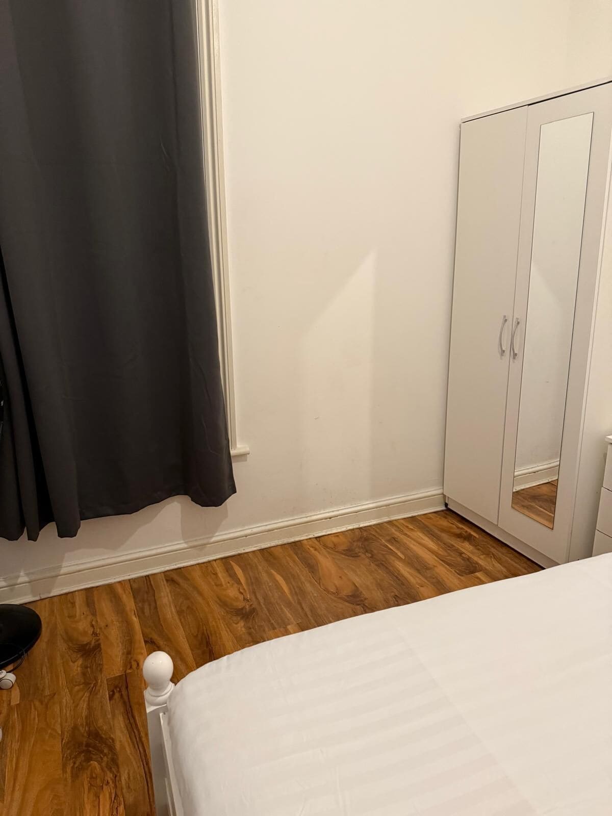 A bedroom with a wooden floor, a white bed, a wardrobe with a mirror, and a window with dark curtains.