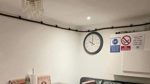 A dining area with a table, chairs, a clock, and a couch. There are signs on the wall indicating no smoking and to keep the kitchen and dining area clean.