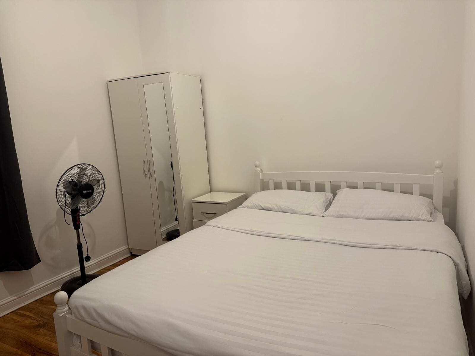 A bedroom with a white bed, a white wardrobe, a bedside table, and a standing fan.