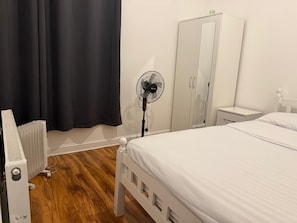 A bedroom with a bed, a radiator, a standing fan, a wardrobe, and a nightstand. - Rayan's Lodge Stratford London by OYO (London)