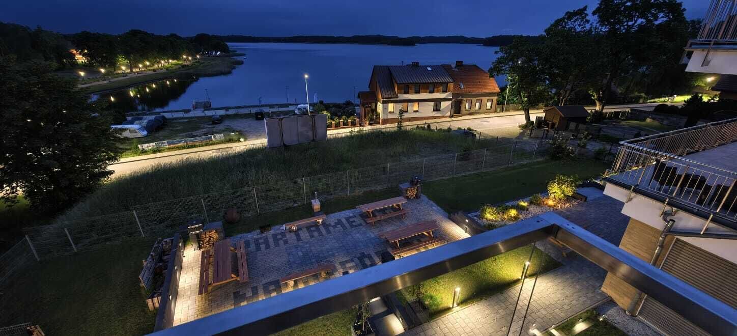 Un edificio moderno con techo plano, un patio empedrado con mobiliario exterior y vistas a un cuerpo de agua.