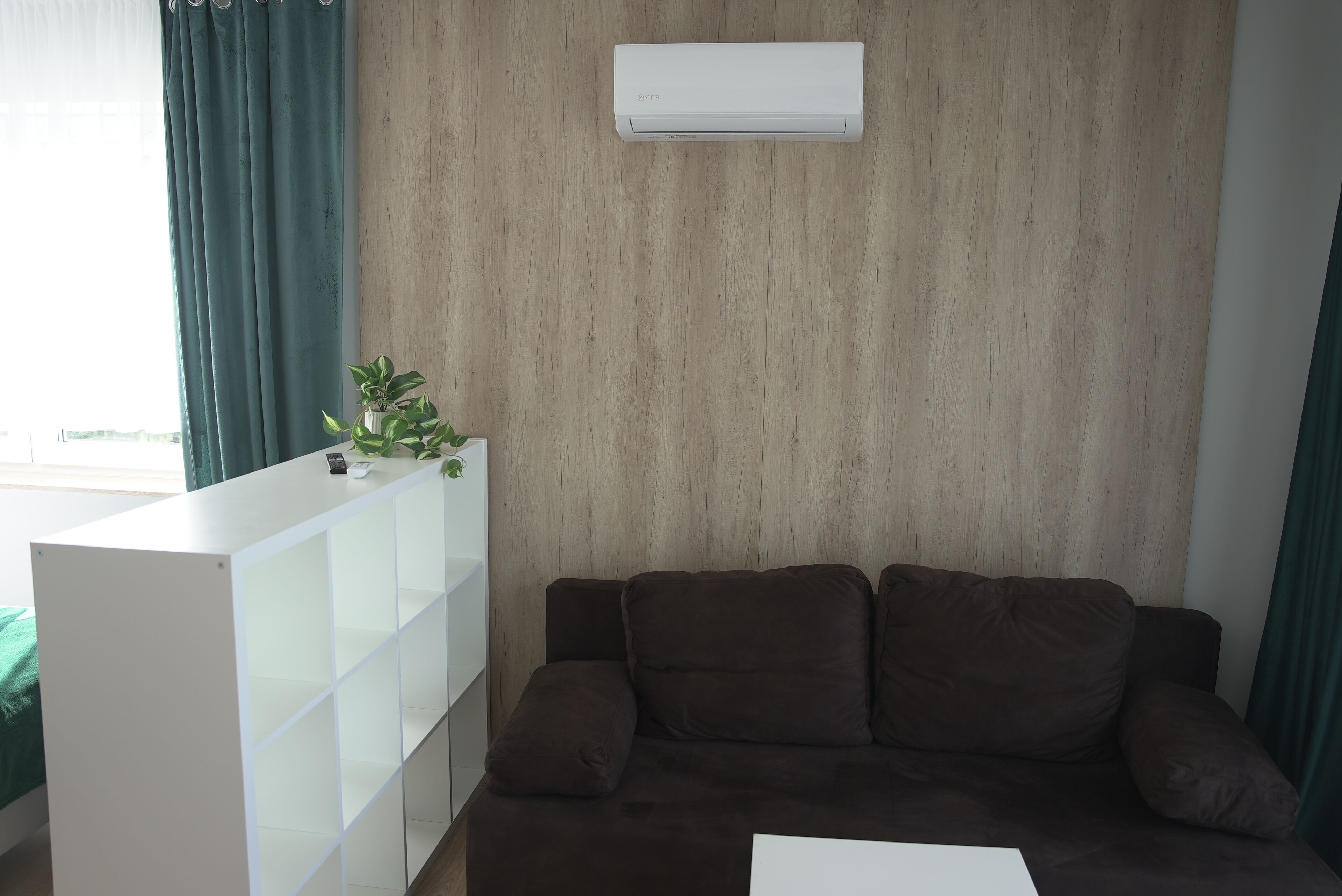 A living room with a brown sofa, a white shelving unit, and a wall-mounted air conditioner.