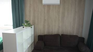 A living room with a brown sofa, a white shelving unit, and a wall-mounted air conditioner.