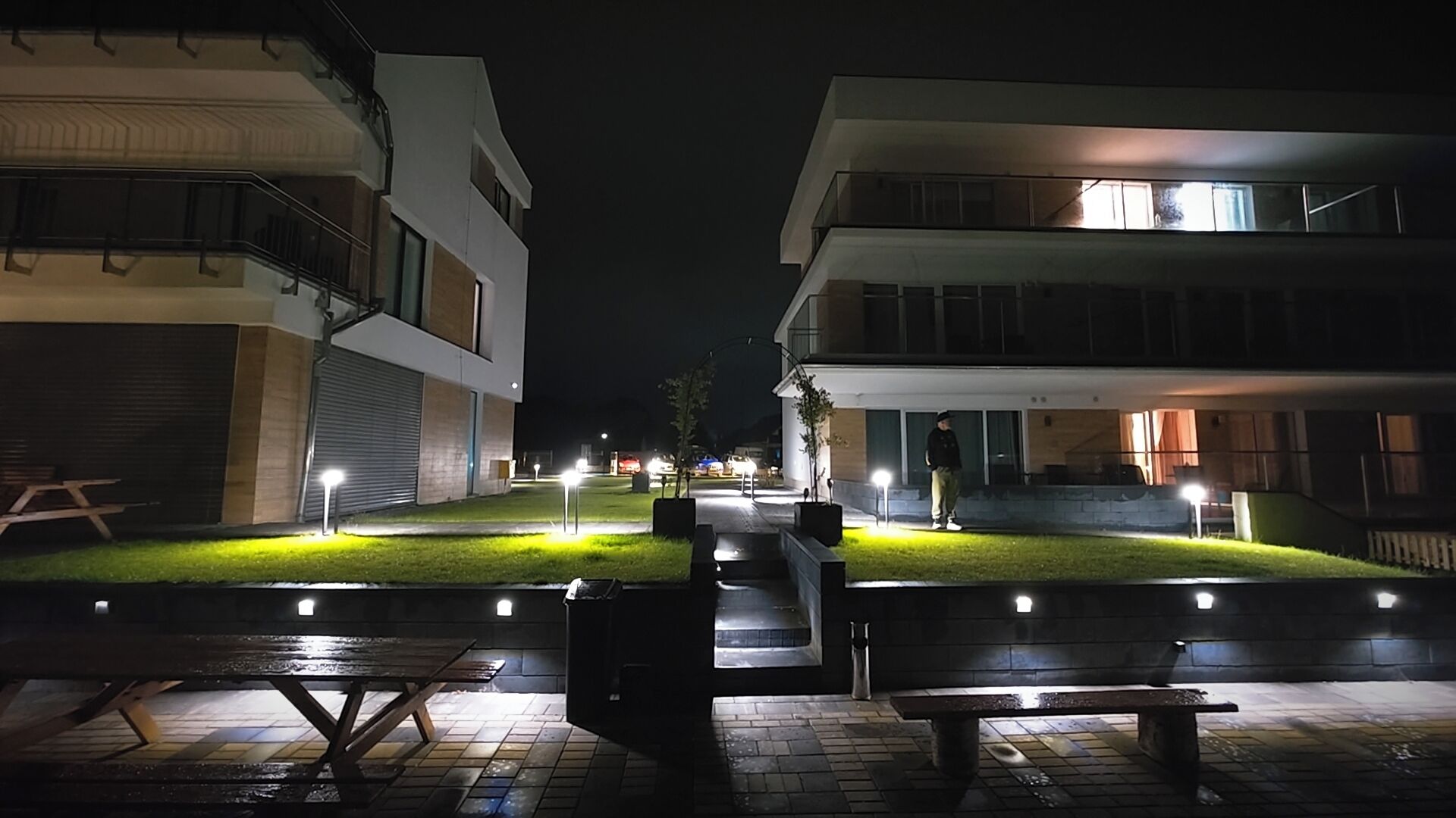 Vista nocturna de un patio con mesas al aire libre y senderos iluminados, rodeado de edificios modernos.