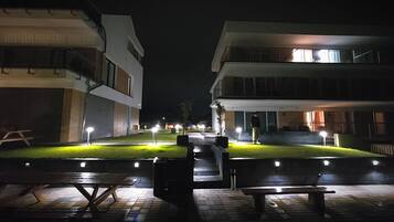 Vista nocturna de un patio con mesas al aire libre y senderos iluminados, rodeado de edificios modernos.