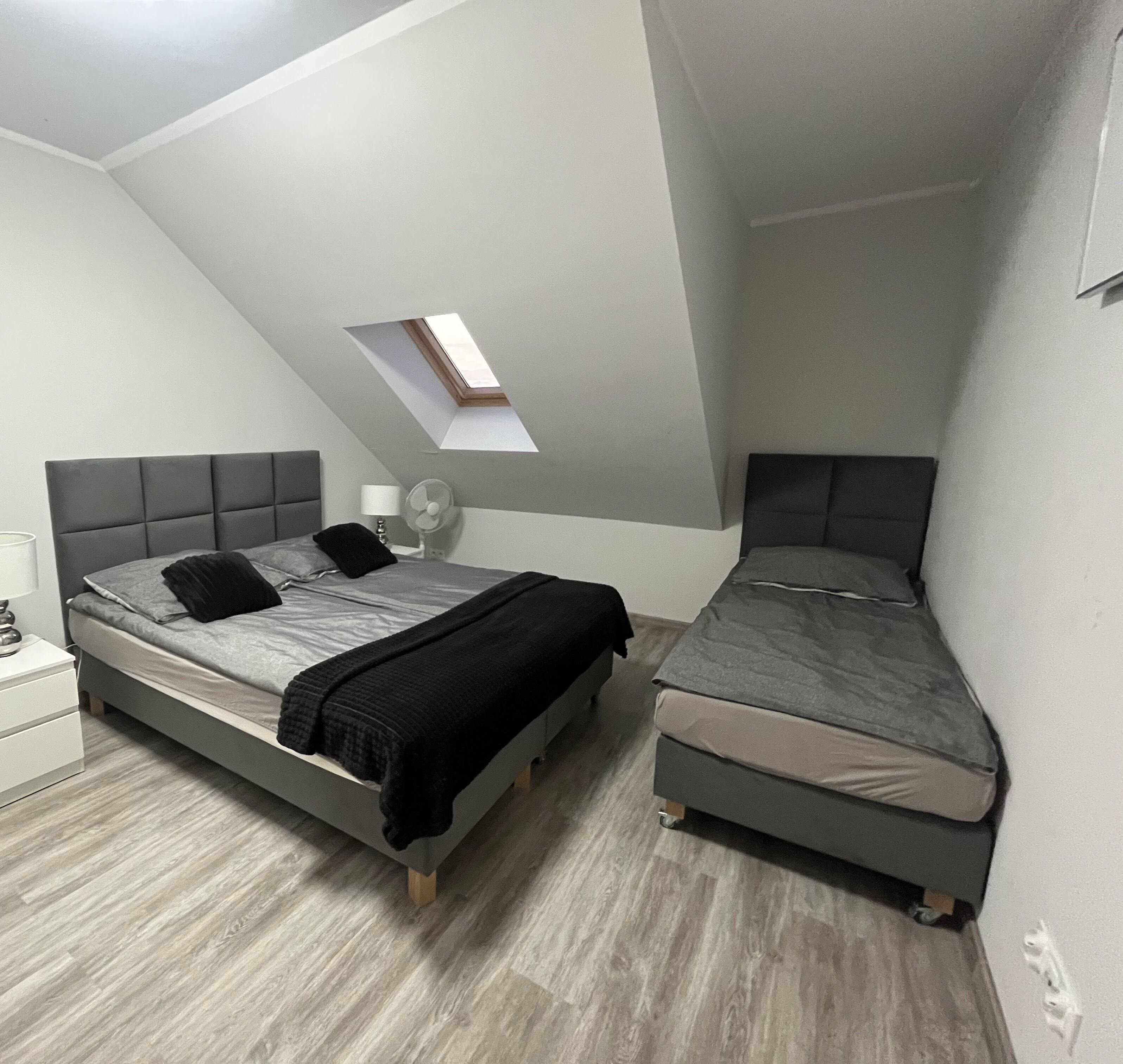 Un dormitorio con cama tipo altillo y una cama más pequeña, tragaluz, mesita de noche y otra mesita auxiliar.