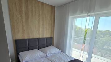 A modern bedroom with a wooden headboard, a large bed with white linens, and a view of a balcony with a cityscape.