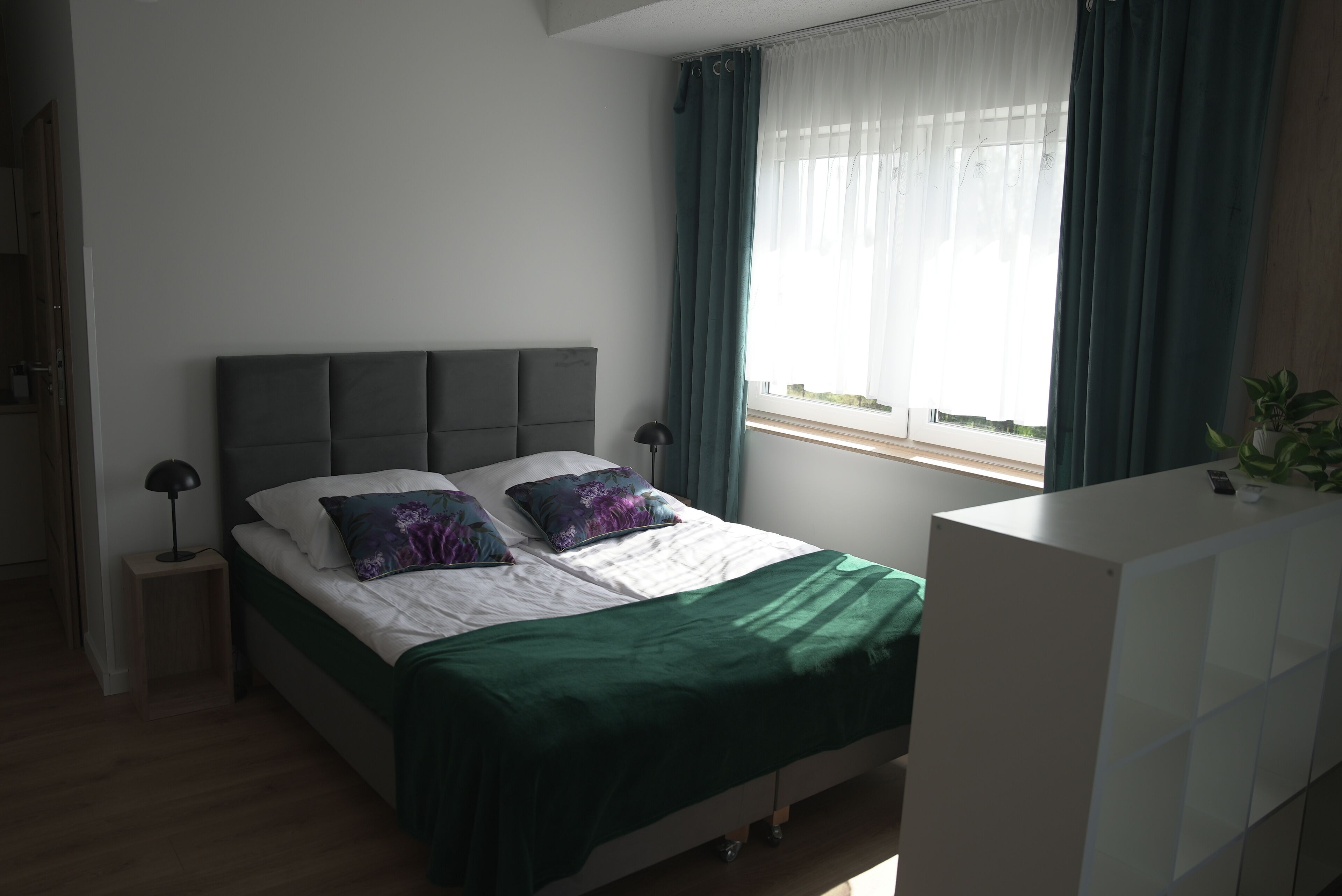 Un dormitorio con cama, mesita de noche, lámpara, ventana con cortinas y una planta.