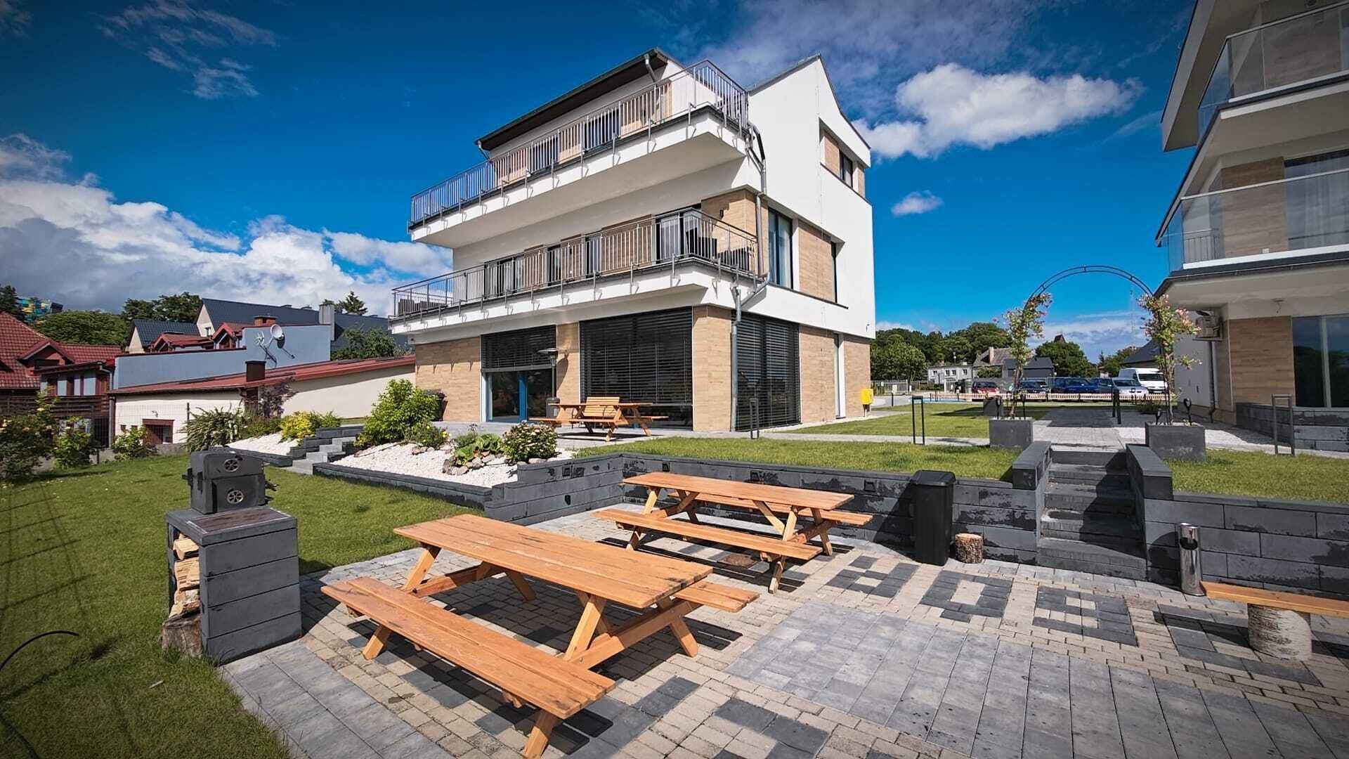 A modern two-story building with a patio area featuring wooden picnic tables and a barbecue grill.