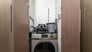 A compact laundry room with a front-loading washing machine, a drying rack, and a large bucket.