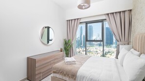 A modern bedroom with a large bed, a bedside mirror, a wooden dresser, and a view of a cityscape through large windows.