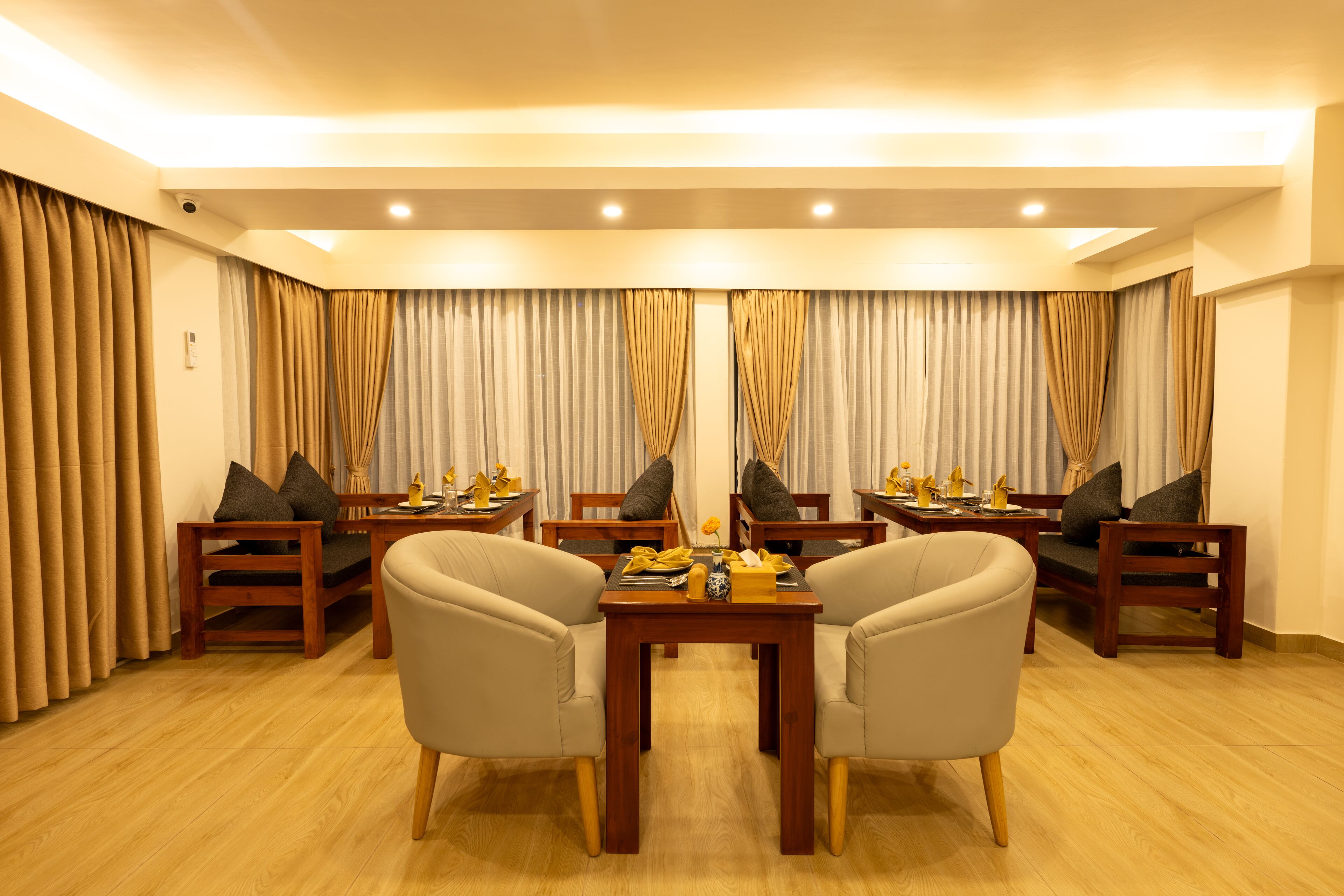 A hotel room with a seating area featuring two armchairs and a small table, wooden flooring, and curtains on the windows.