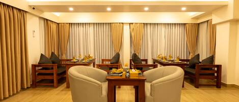 A hotel room with a seating area featuring two armchairs and a small table, wooden flooring, and curtains on the windows.