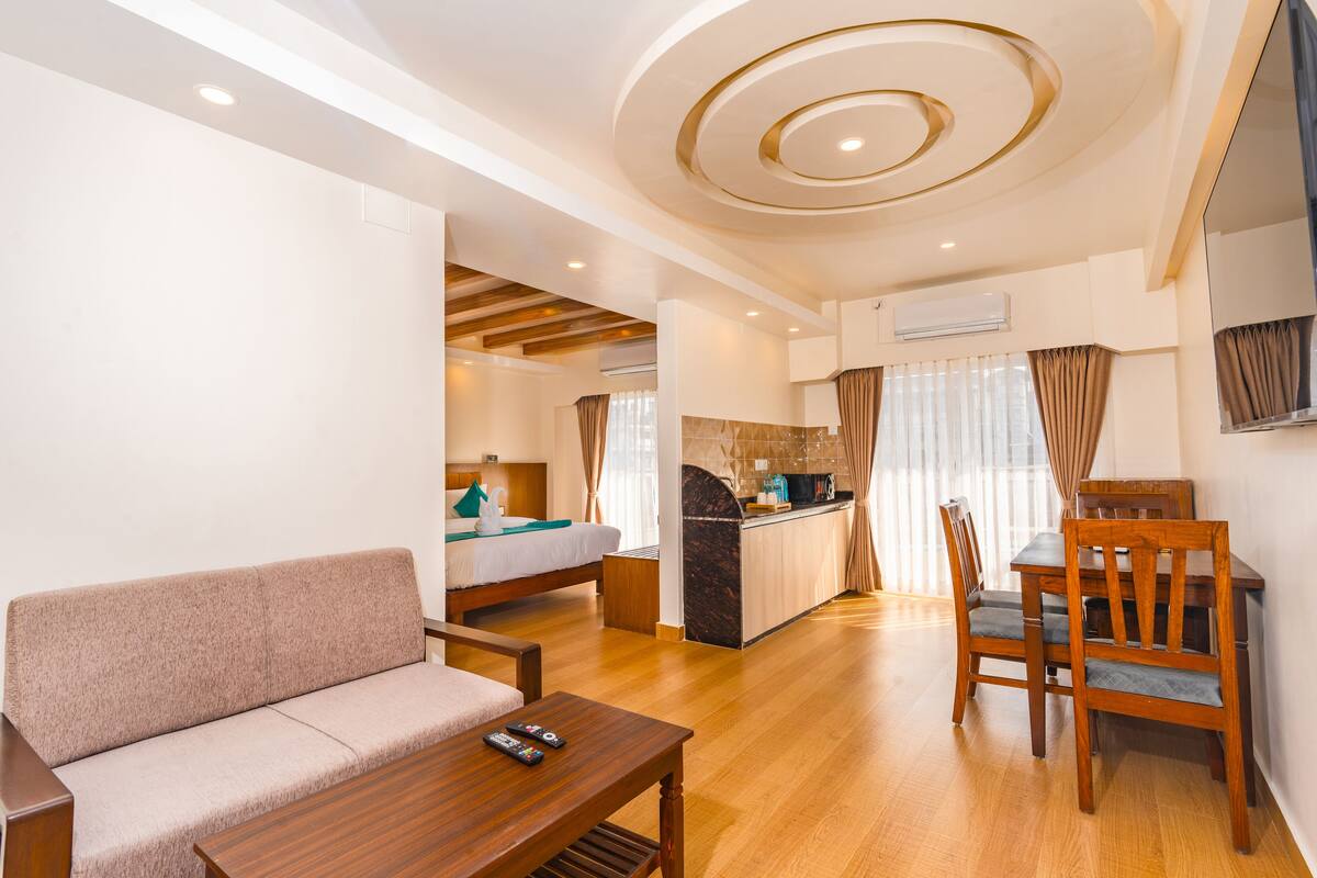 A hotel room with a sofa, a wooden table, a dining table with chairs, a kitchen area, and a ceiling with circular light fixtures.