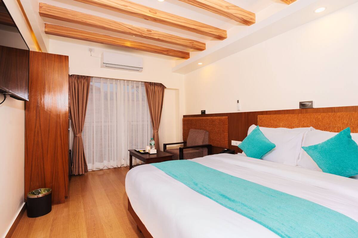 A hotel room with a large bed, wooden ceiling beams, a desk with a chair, and a small trash bin.