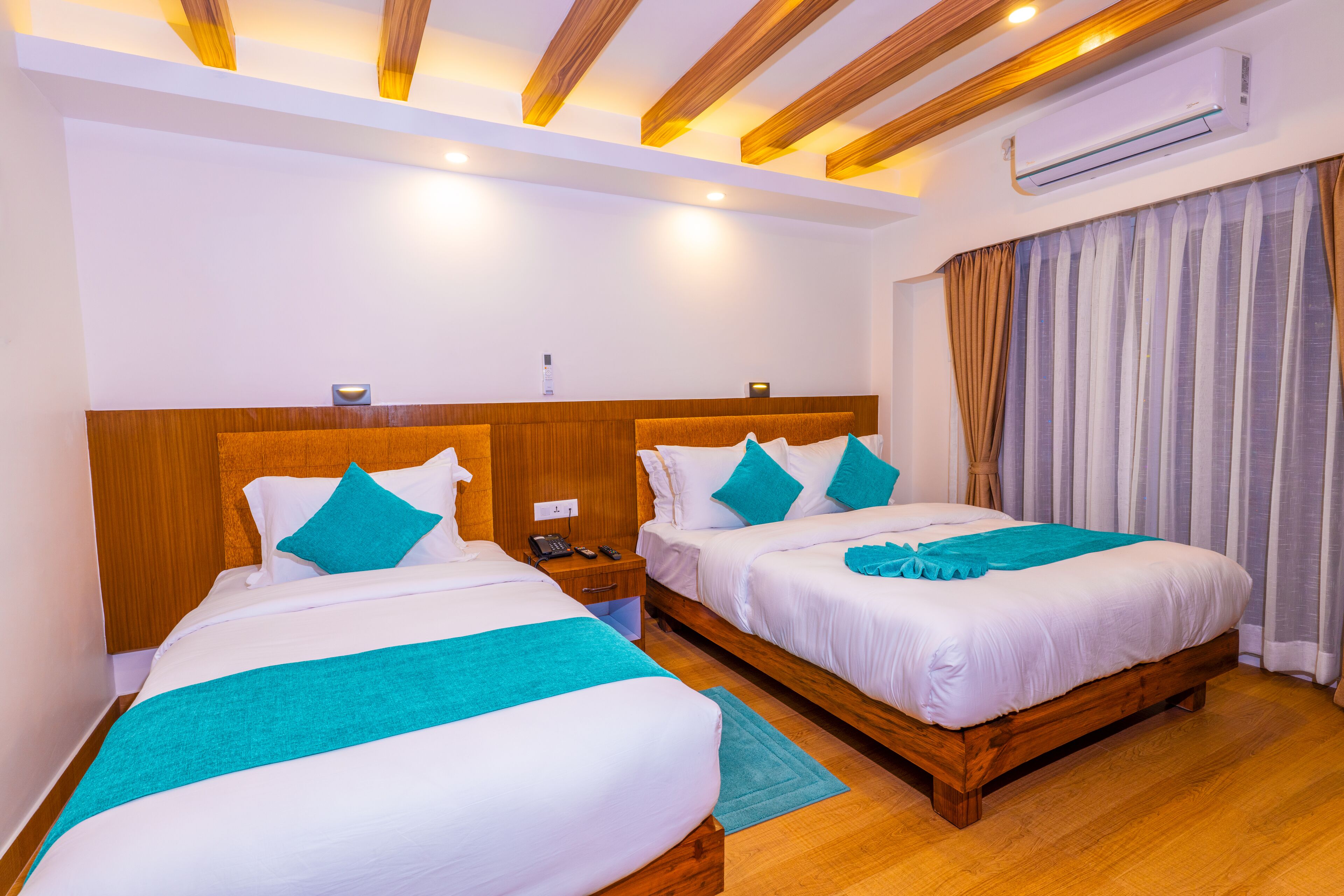 A hotel room with two beds, wooden headboards, white bedding, teal throw pillows, a wooden floor, an air conditioning unit, and a nightstand with a phone.