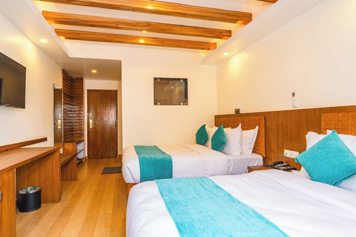 A hotel room with two beds, wooden headboards, a desk, a TV, and a small window.