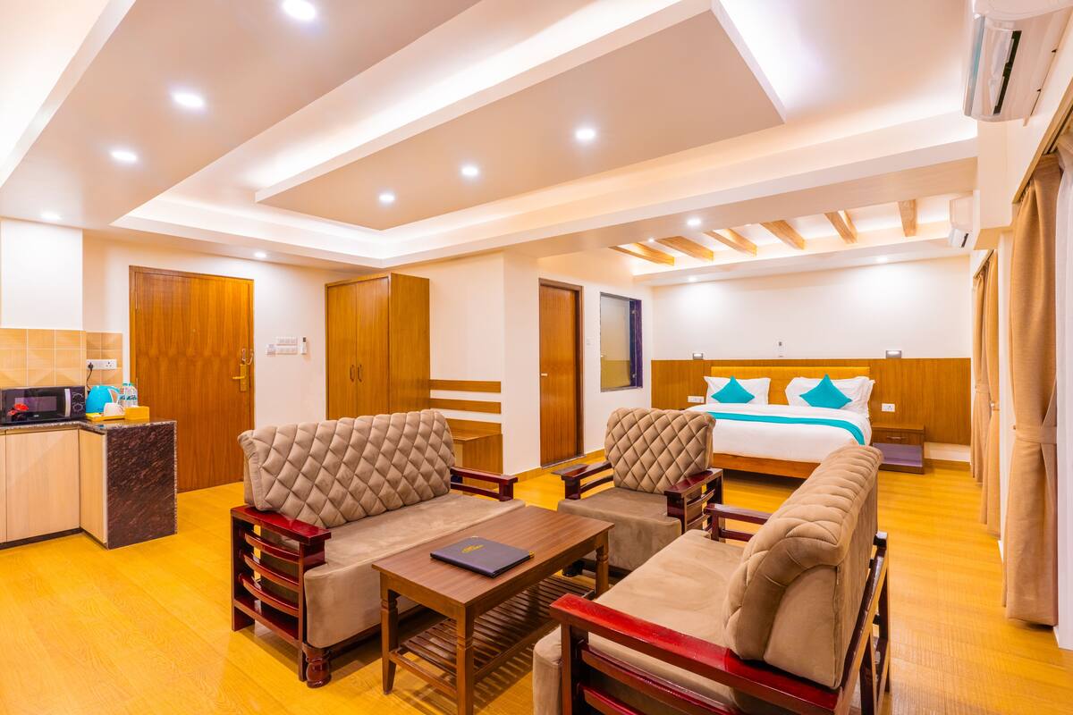 A modern hotel room with a sofa set, a dining table, a bed with a blue headboard, and a kitchen area with a microwave.