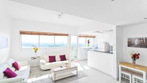 Ein geräumiger Wohnbereich mit einem weißen Sofa, einem Couchtisch und einem Esstisch mit Stühlen. Große Fenster bieten Blick auf die Stadt.