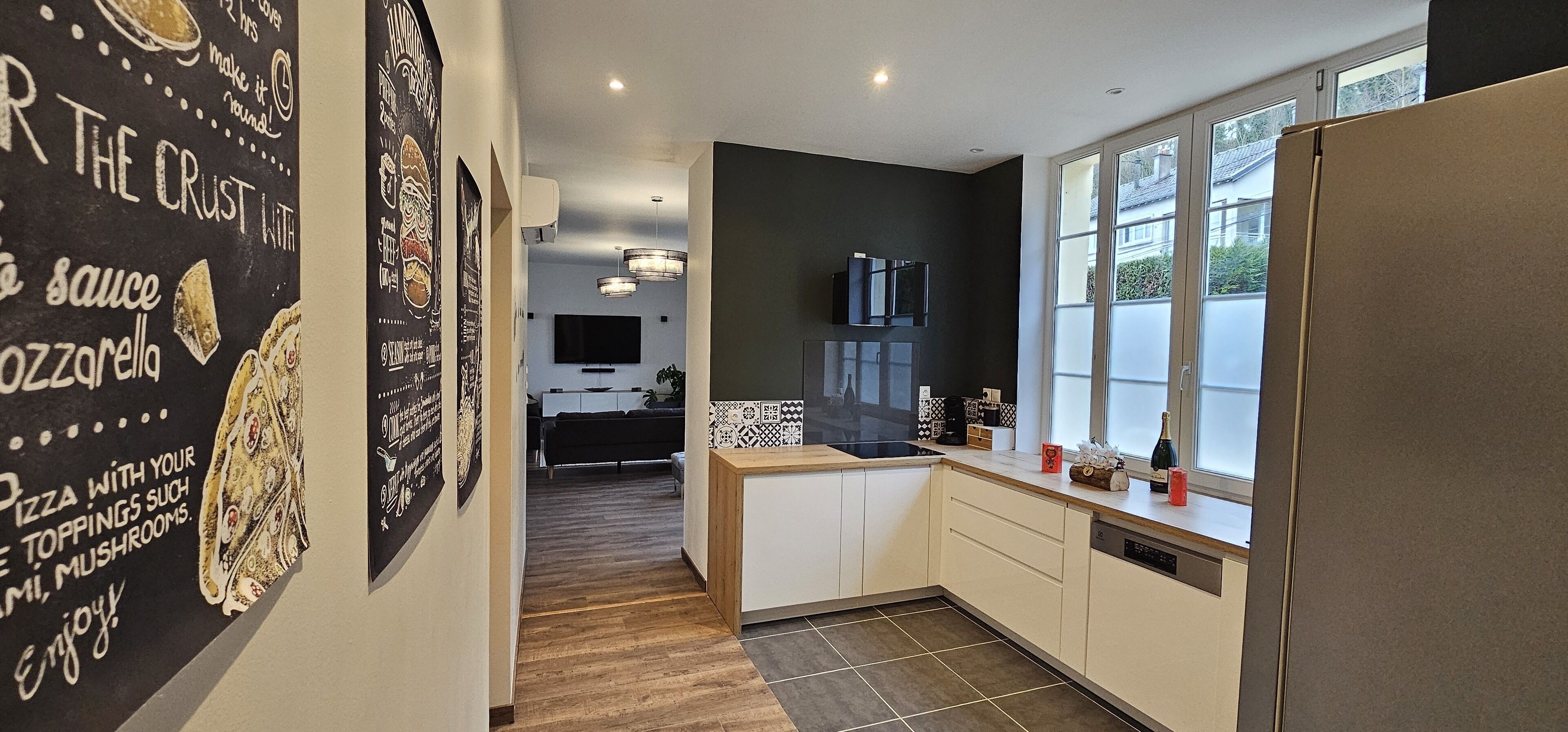 A modern kitchen with a wooden floor, a large window, and a refrigerator.