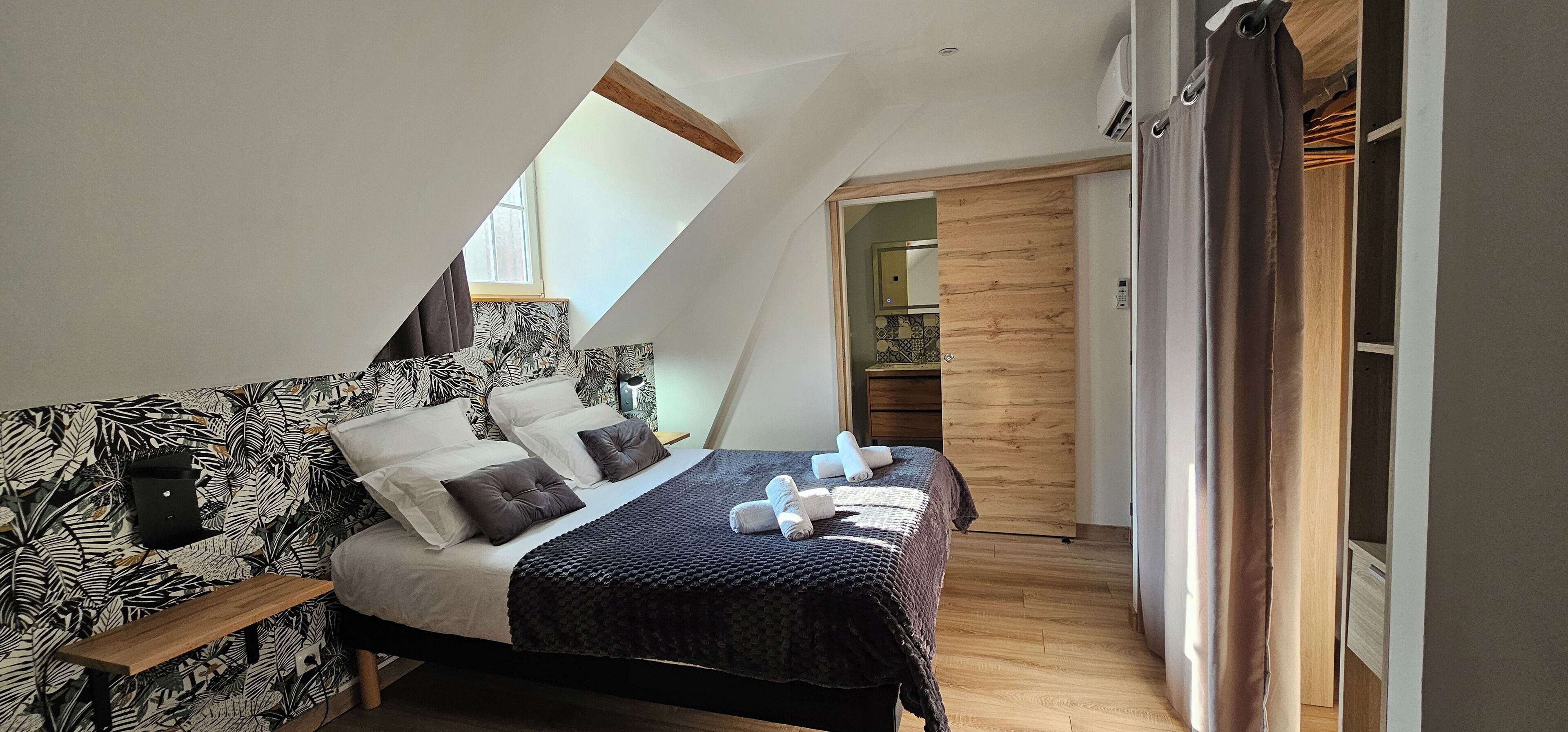 A bedroom with a sloped ceiling, a bed with a patterned headboard, a wooden bench, and a view of a kitchen through an open door.