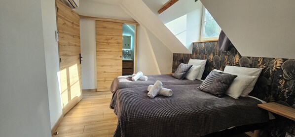 A bedroom with two beds, wooden flooring, a skylight, and a view of a bathroom.