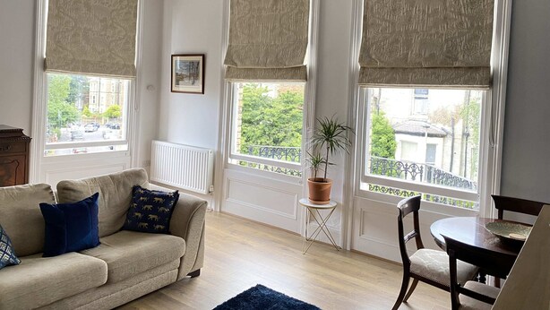 Een woonkamer met een beige bank, een klein rond tafeltje en een stoel. Er zijn grote ramen met beige gordijnen, een radiator en een bloempot op een klein tafeltje.