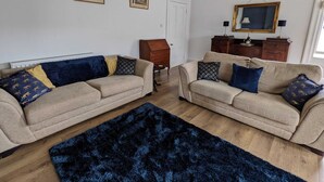 Een woonkamer met twee beige banken, een donkerblauwe vloerkleed, houten meubels en lijstwerken aan de muur.