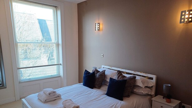 Een net opgemaakt bed met witte lakens en opgevouwen handdoeken, een nachtkastje met een lamp, en een raam met zonwering.