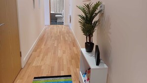 Een gang met houten vloer, een potplant, een televisie op een witte console en een kleurrijk vloerkleed.