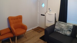 Et værelse med trægulv, en sort sofa med et ternet pudebetræk, en orange lænestol og en hvid dør med et vindue.