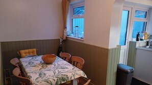 Et spiseområde med et bord dækket til fire personer, et vindue med gardiner, et lille køkkenområde med en vask og en skraldespand, samt træstole med hynder.