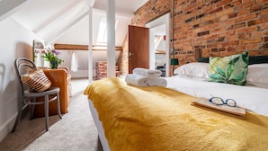 Hotellihuone, jossa on tiiliseinä, keltainen peitto sängyllä, punottu kassi tuolissa ja ikkuna, josta avautuu näkymä parvelle.