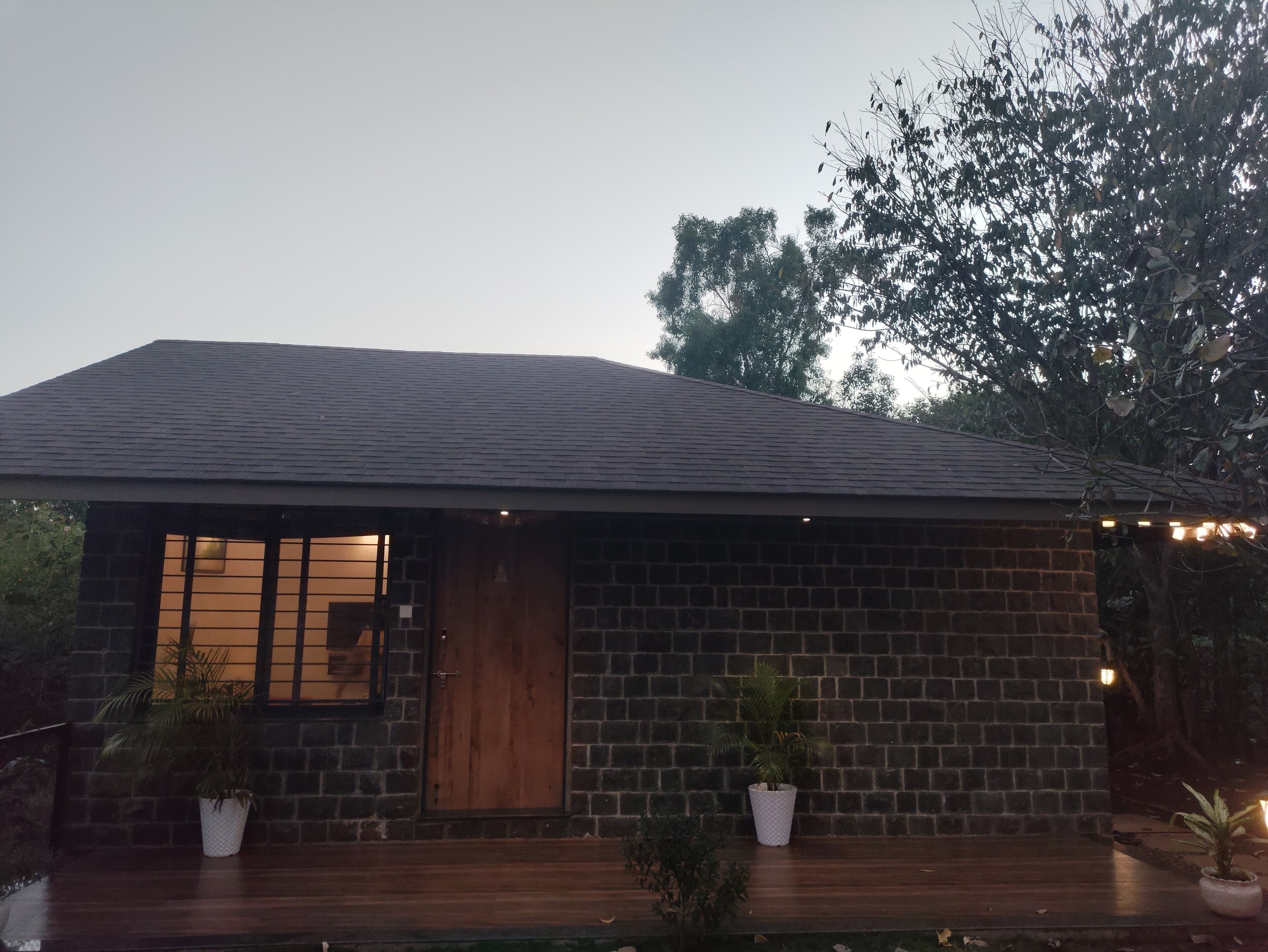 Een bakstenen huis met een houten deur en ramen, omringd door plantenpotten en bomen.