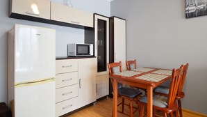 A compact kitchen with a refrigerator, microwave, and cabinets. There is a dining table with chairs and a painting on the wall. - RentPlanet - Apartament Bałtycka (Kolobrzeg)