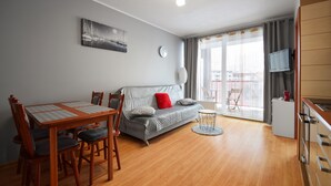 A modern living room with a grey sofa, wooden dining table, and chairs. There is a TV mounted on the wall, a small round table, and a window with curtains. - RentPlanet - Apartament Bałtycka (Kolobrzeg)