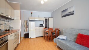 A compact kitchen with modern appliances, a dining area with wooden chairs, and a grey sofa with a red pillow. - RentPlanet - Apartament Bałtycka (Kolobrzeg)