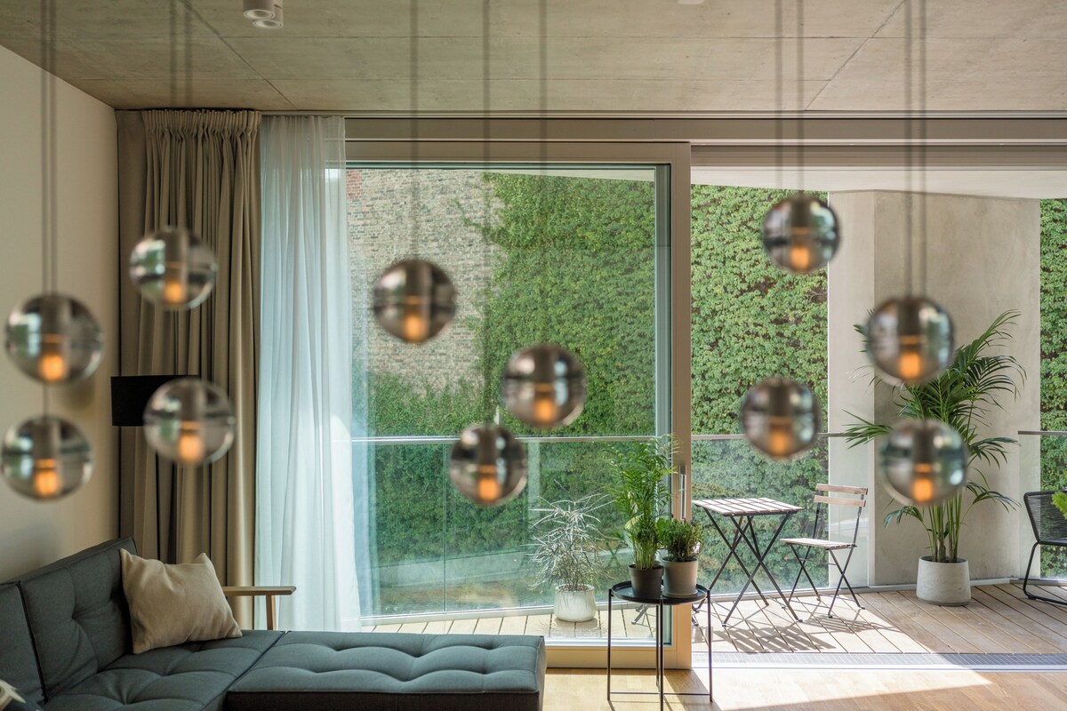 A modern living room with a sofa, a dining table, and chairs, a large window with curtains, and hanging spherical light fixtures.