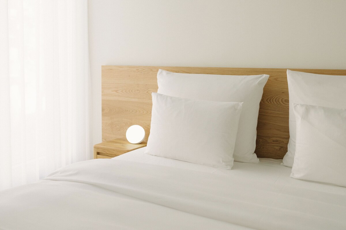 A neatly made bed with white bedding and pillows, a wooden headboard, and a bedside table with a round light.