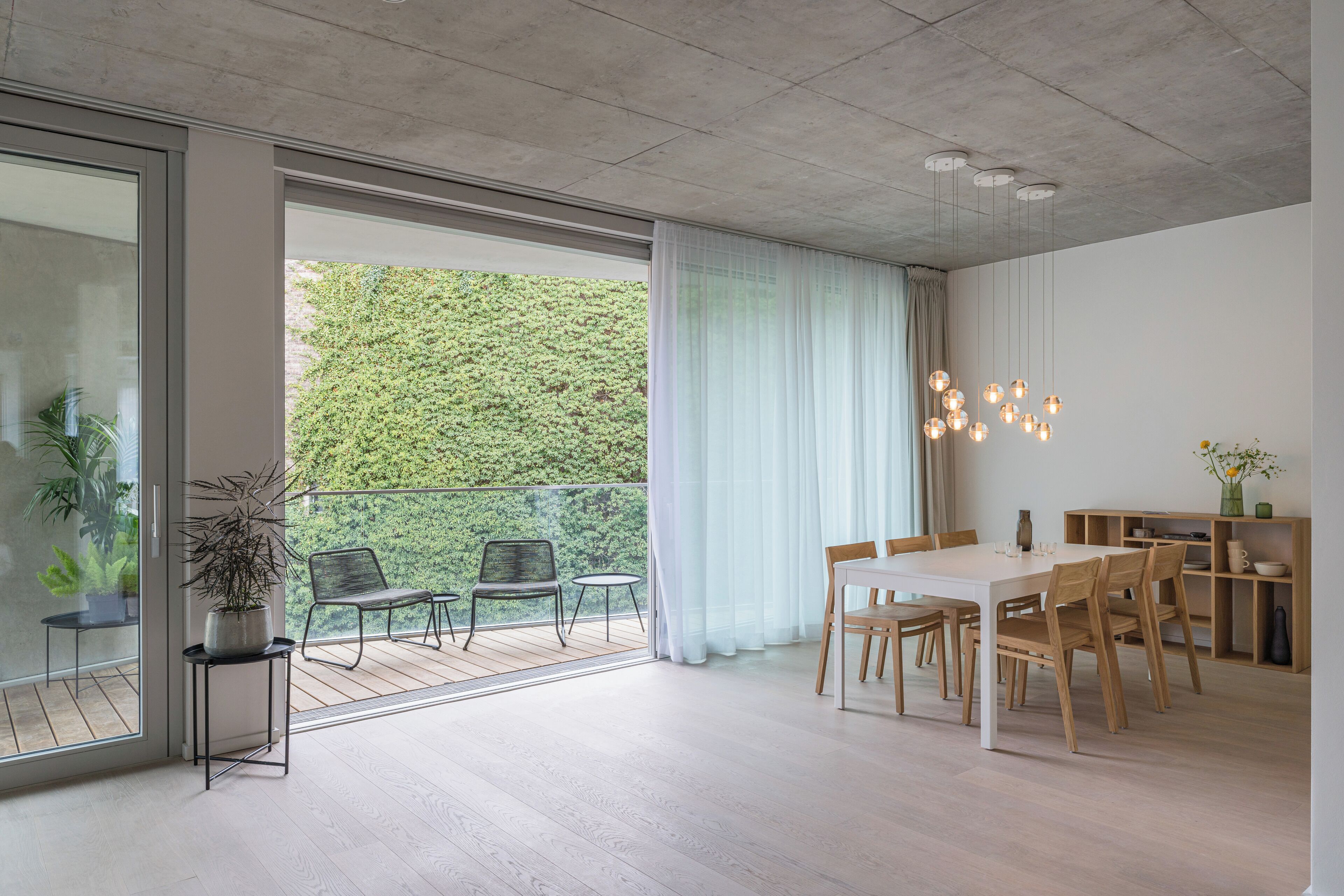 A modern dining area with a large table and chairs, a balcony with chairs, and a potted plant.