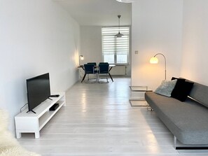 A modern living room with a flat-screen TV, a sofa, a dining area with chairs, and a large window with blinds.